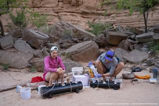 Two people wearing outdoorsy clothes and baseball caps are sitting on the sound surrounded by scientific equipment. They're pouring water into filters attached by plastic tubing to vacuum pumps. 
